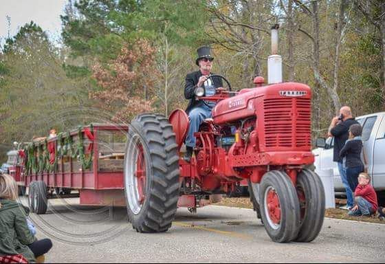Red vintage tractor pulling a decorated trailer at an outdoor parade, with people watching nearby