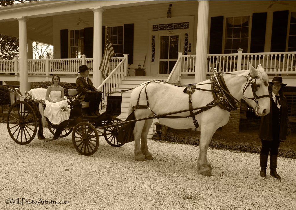 Horse-drawn carriage parked before a porch-lined building, with two people seated and a driver standing beside the horses.
