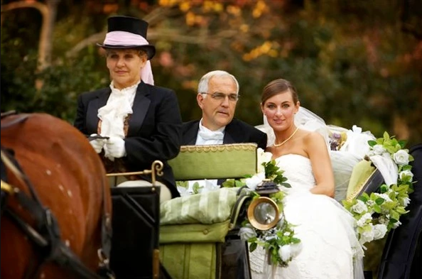 Bride and groom in a horse-drawn carriage with a driver, outdoors in fall colors