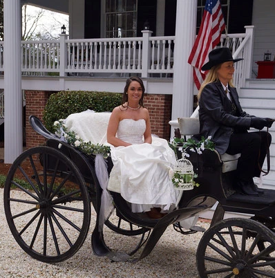 Bride in white dress seated in a horse-drawn carriage outside a house with an American flag