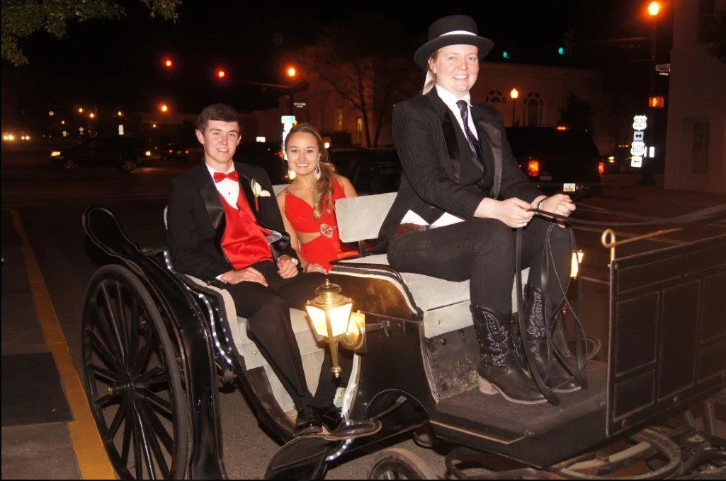 Two people in formal attire riding in a horse-drawn carriage at night, lit by streetlights.