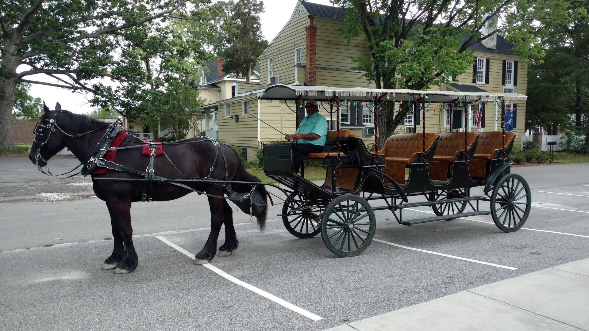 Horse-drawn carriage with passengers crossing a street in front of houses and trees.