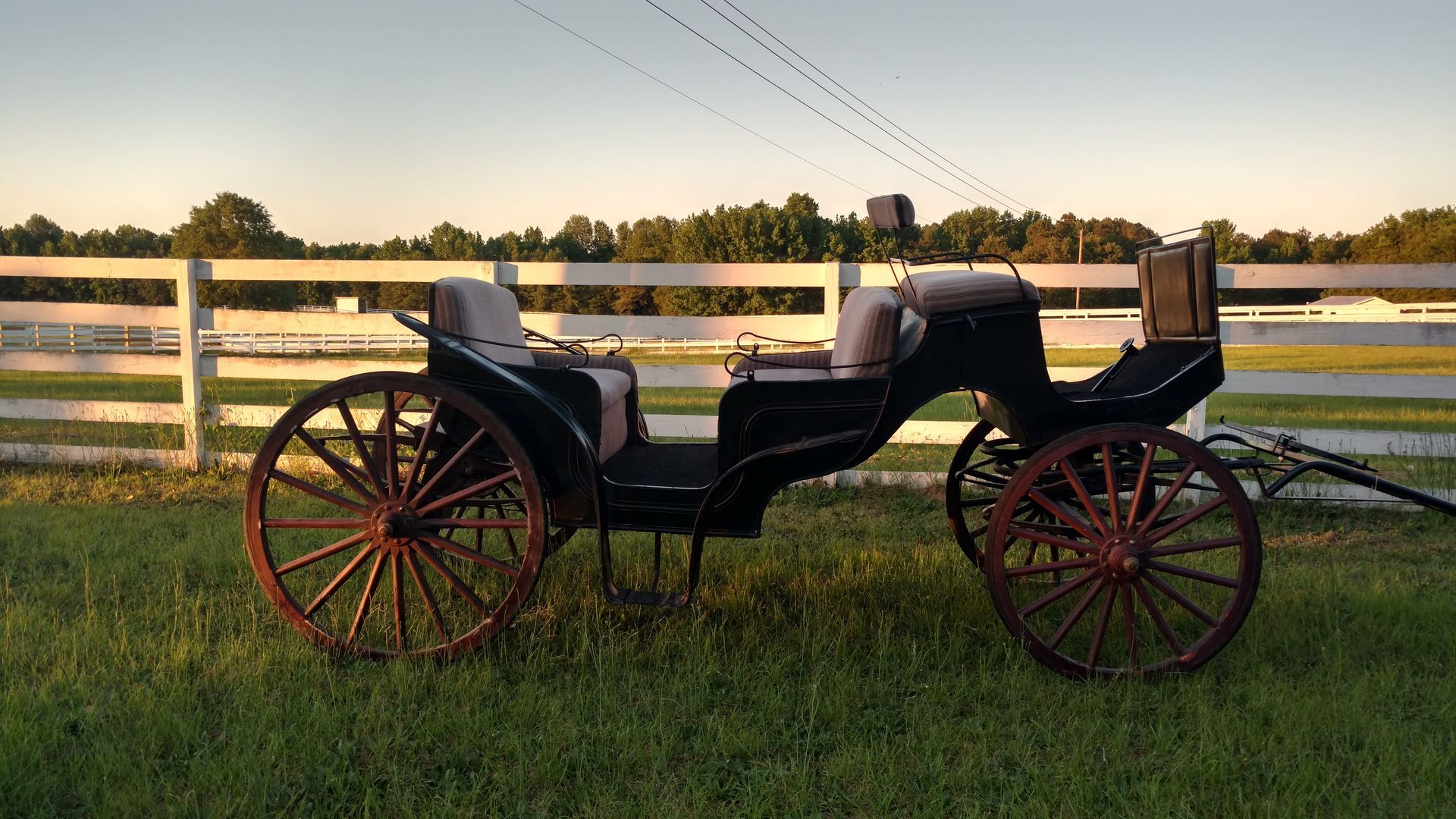 Vintage horse-drawn carriage on grass by a white fence at sunset