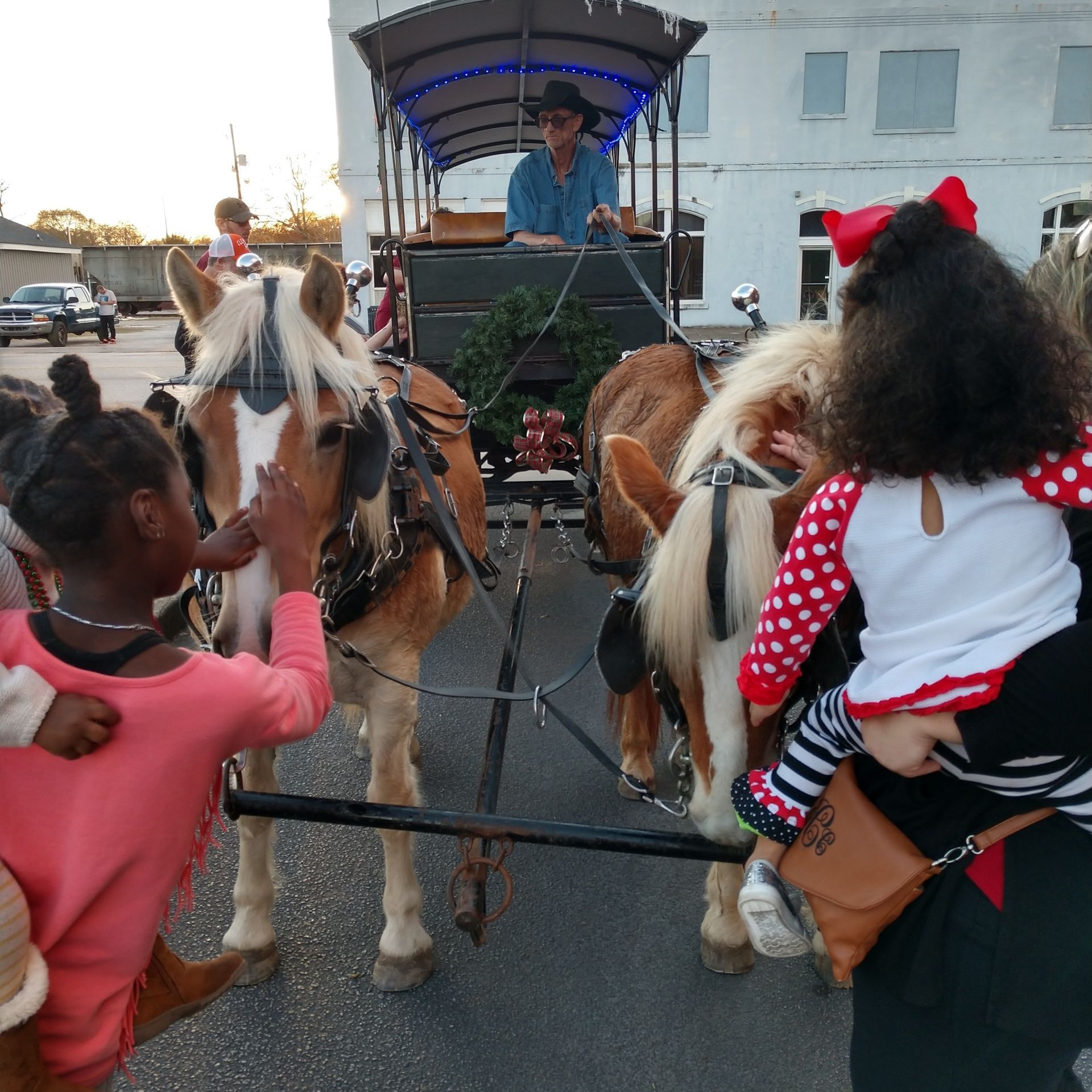 Children riding ponies on a carriage trail, with a driver under a blue canopy in a parking lot.