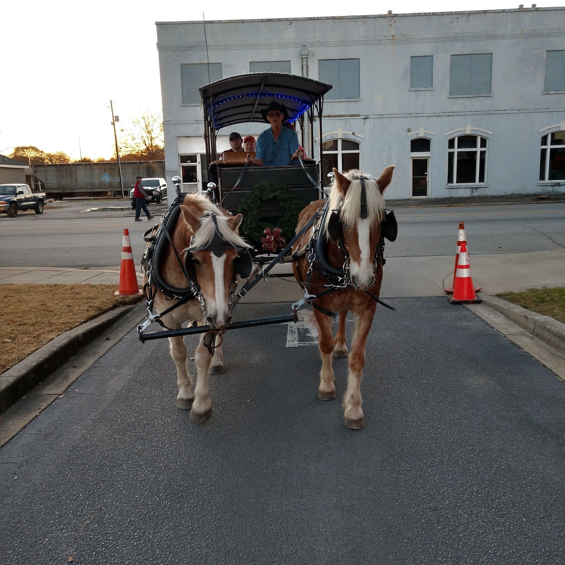 Two horses pulling a carriage down a street past orange cones and a white building at dusk