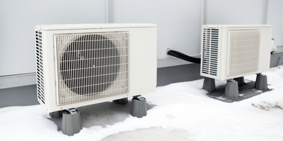 Two white outdoor AC units elevated on gray blocks in snow