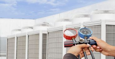 Hands holding red and blue pressure gauges in front of rooftop HVAC units