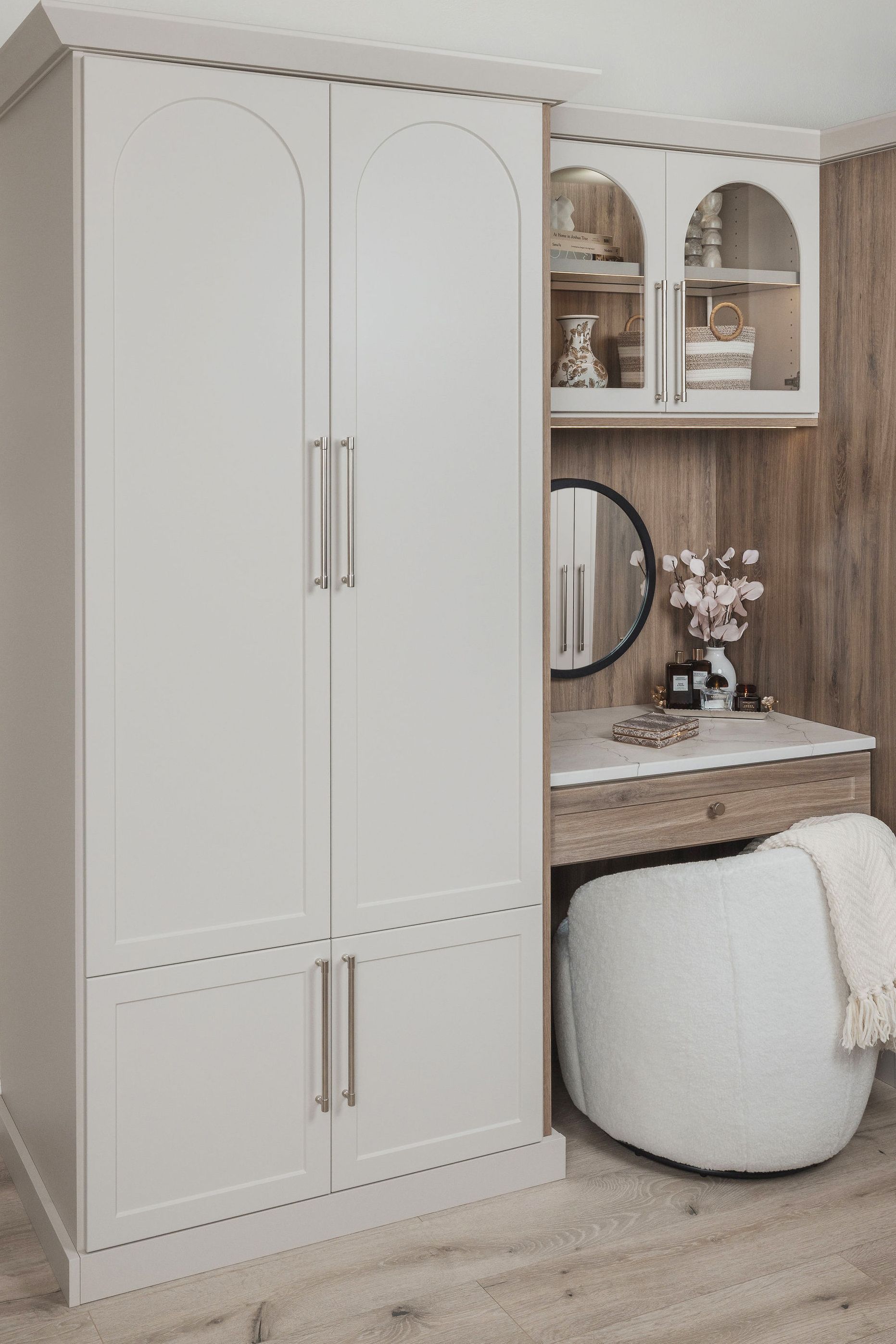 Built-in white wardrobe beside a vanity desk with arched shelves and a round pouf in a beige bedroom.