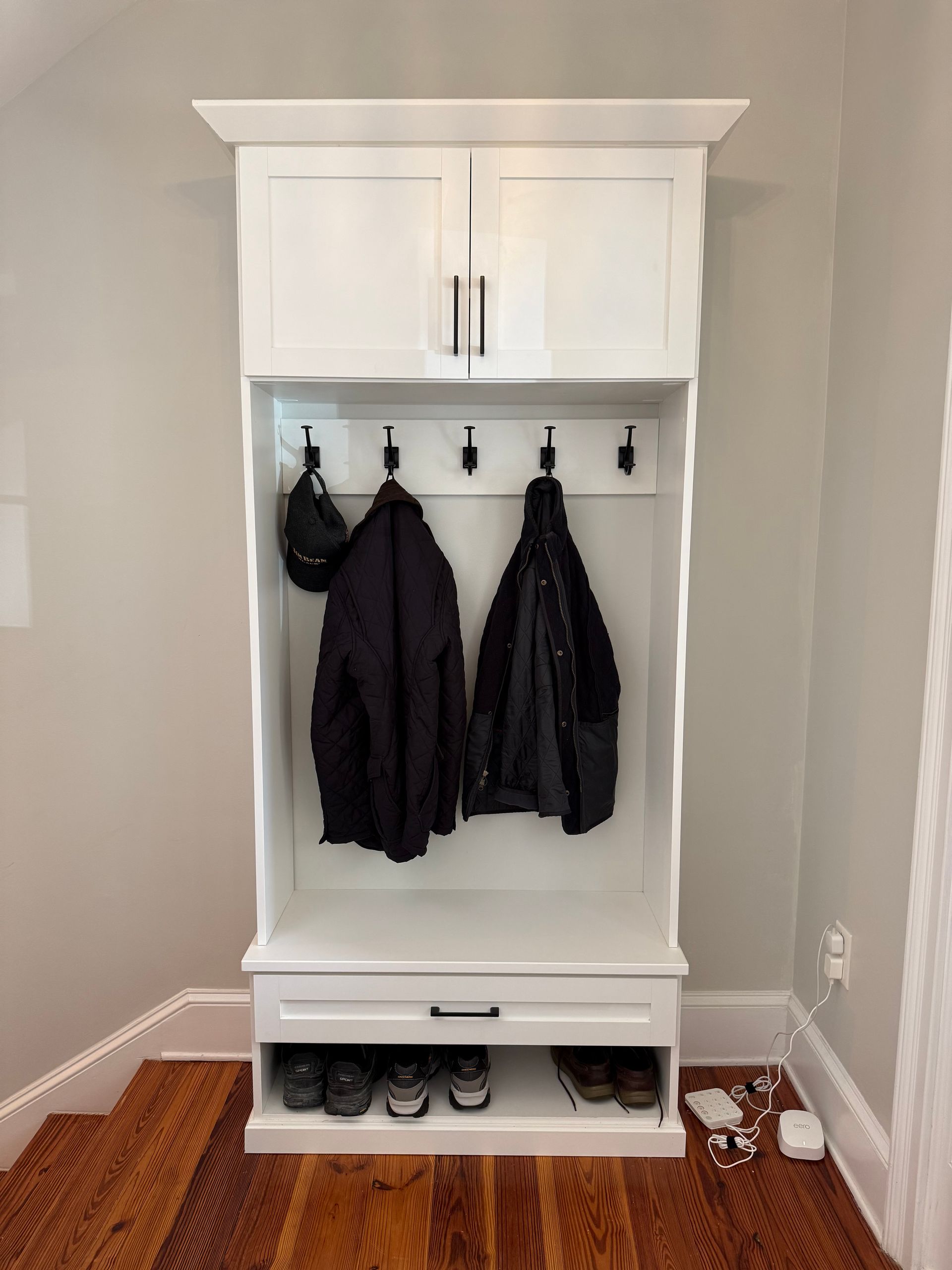 White entryway organizer with hooks, a drawer, and shoe storage; coats and a helmet hang from hooks.