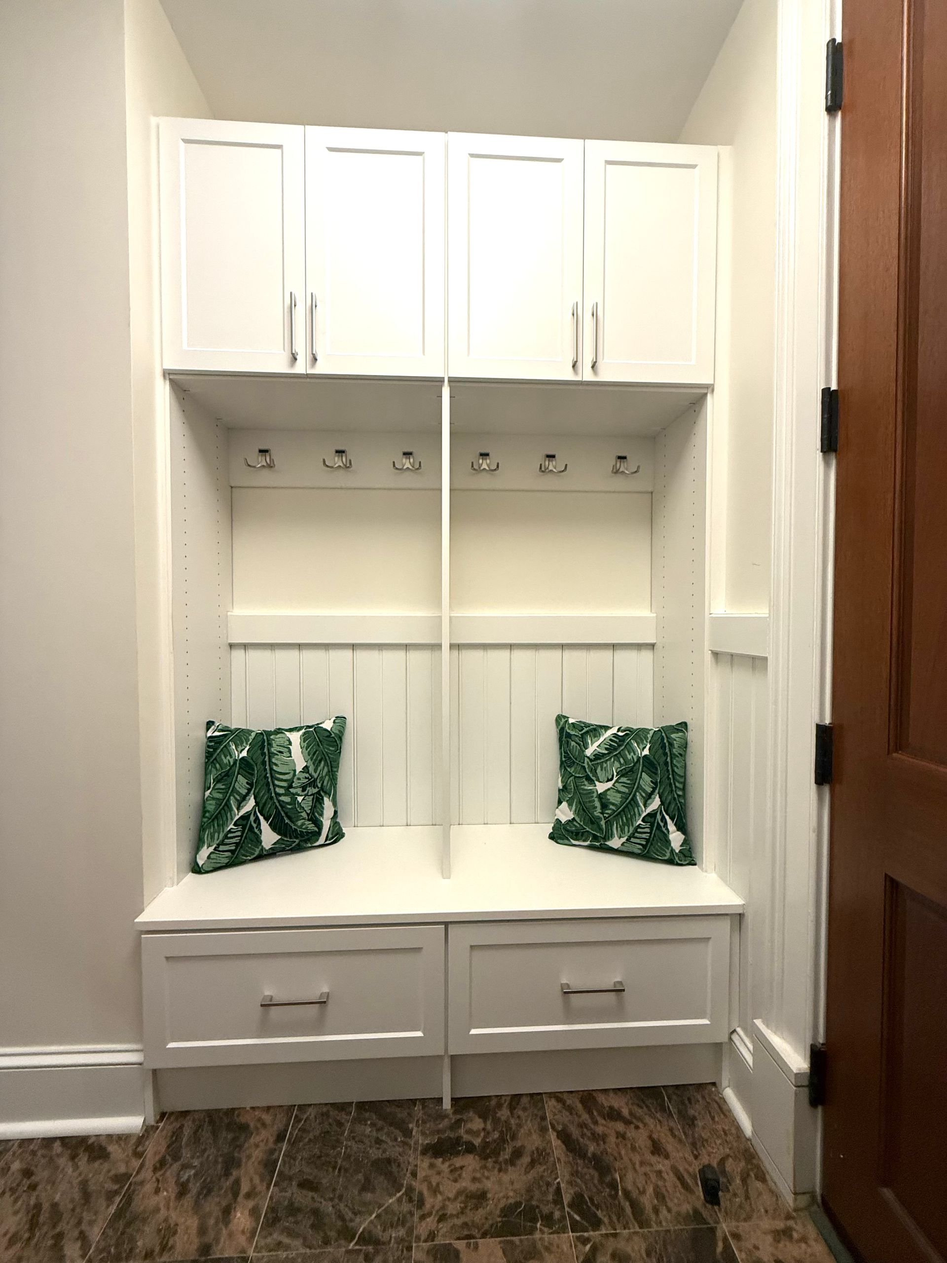White built-in storage unit with cabinets, hooks, and drawers. Two green patterned pillows on the bench.