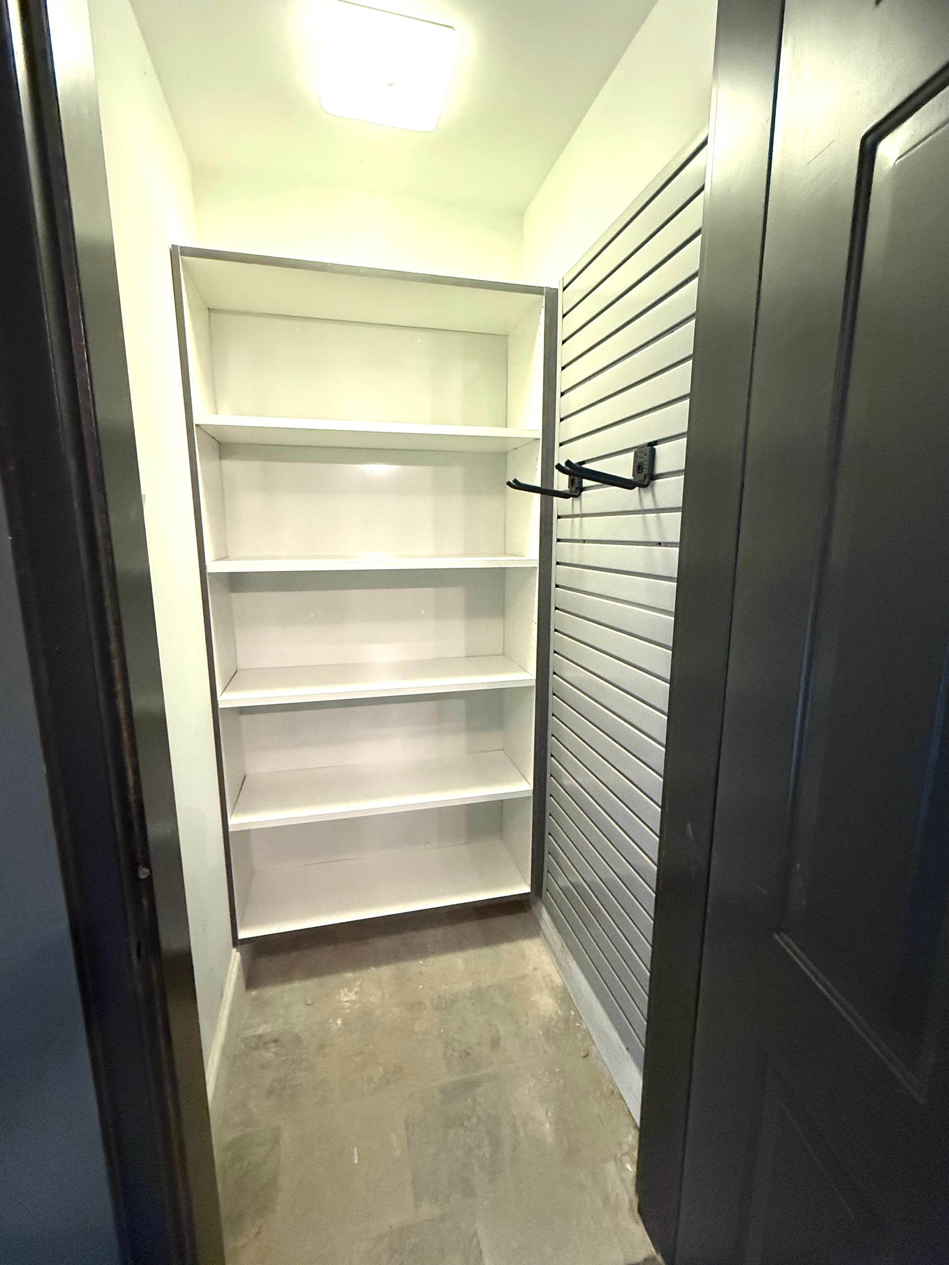 Interior view of a small closet with white shelves and a black door. Wall with metal paneling.