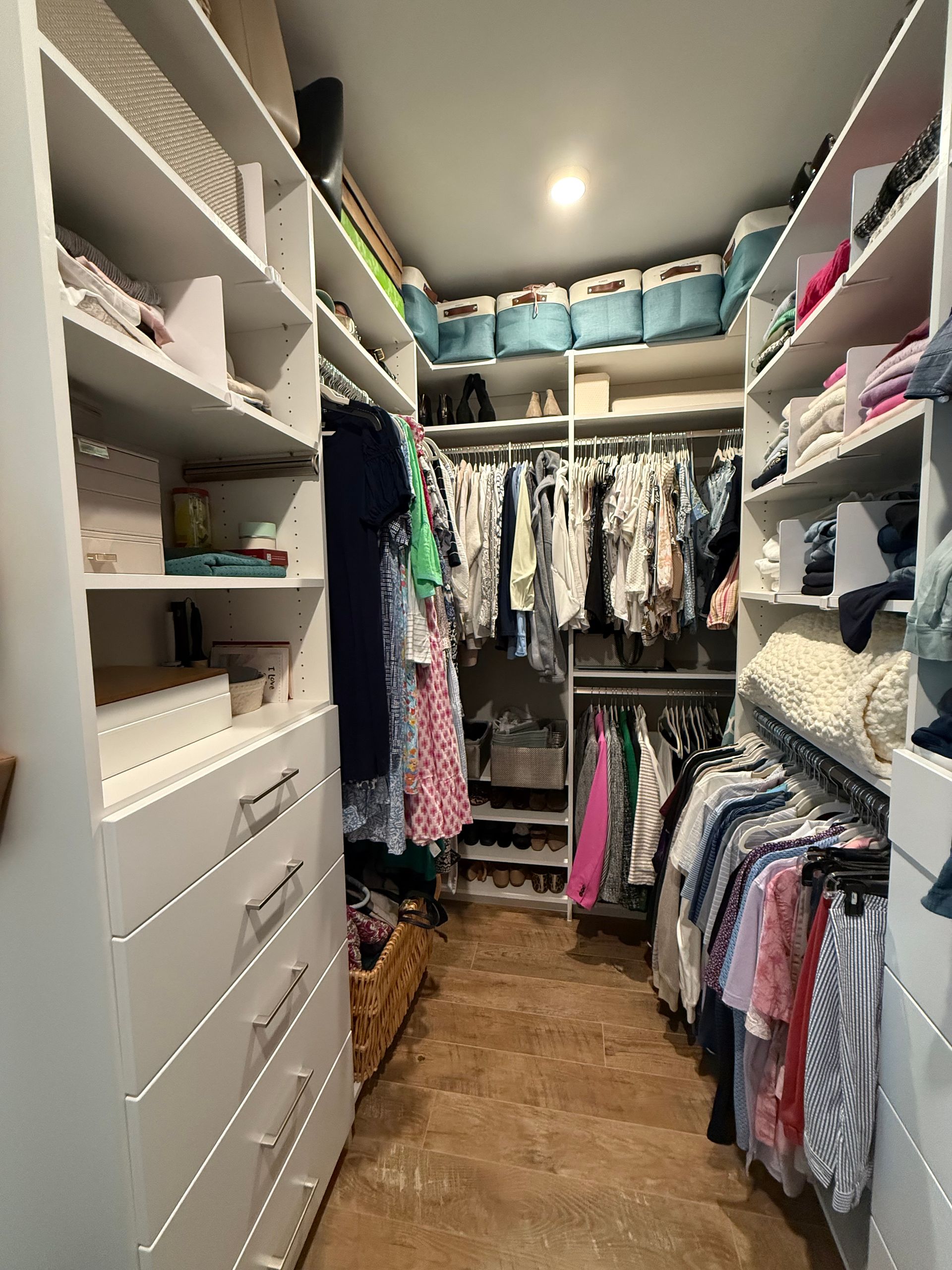 Walk-in closet with white shelving, drawers, and hanging clothes; baskets and storage bins above.