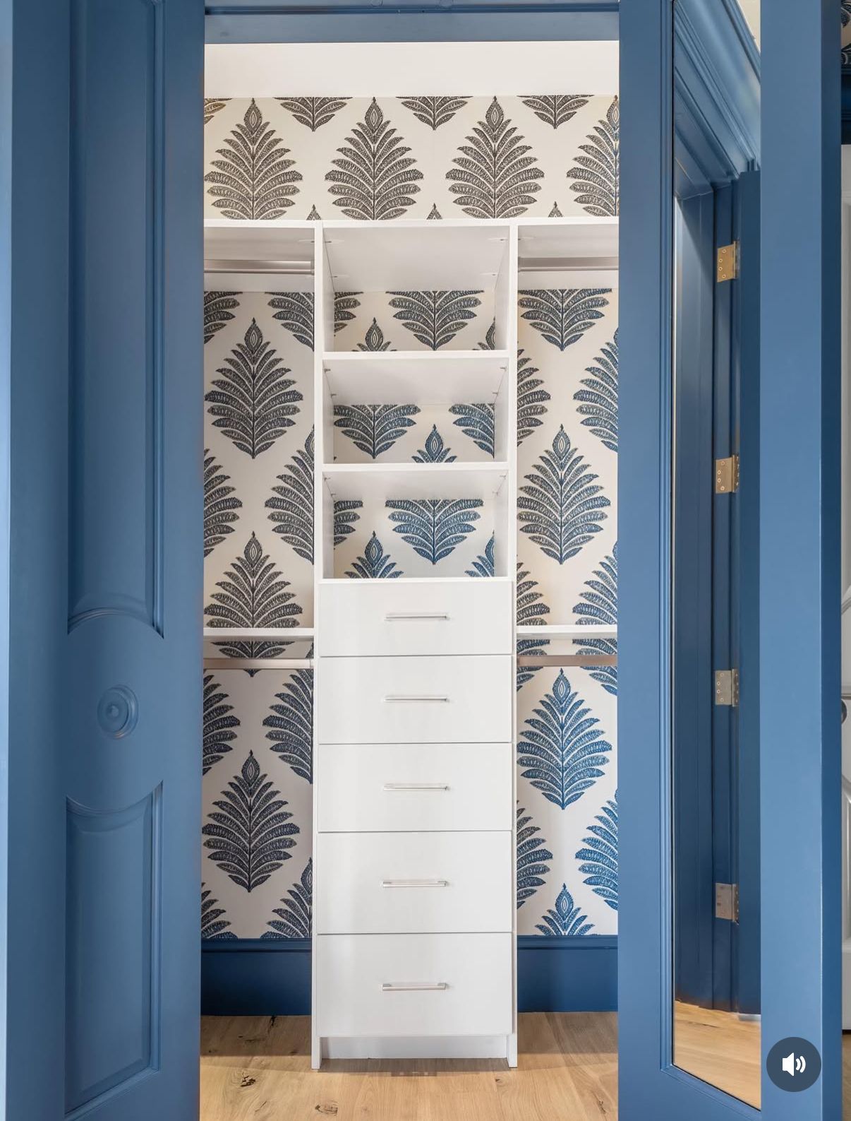 Blue closet doors open to a white closet organizer with drawers, set against patterned wallpaper.
