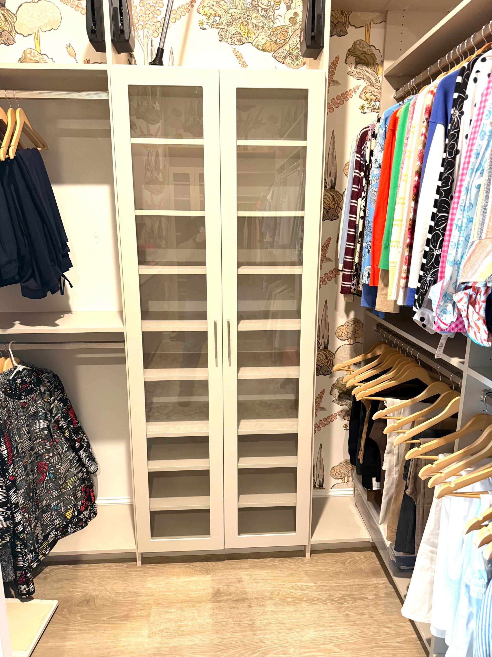 Closet with a tall white cabinet with glass doors; clothes hung on either side.