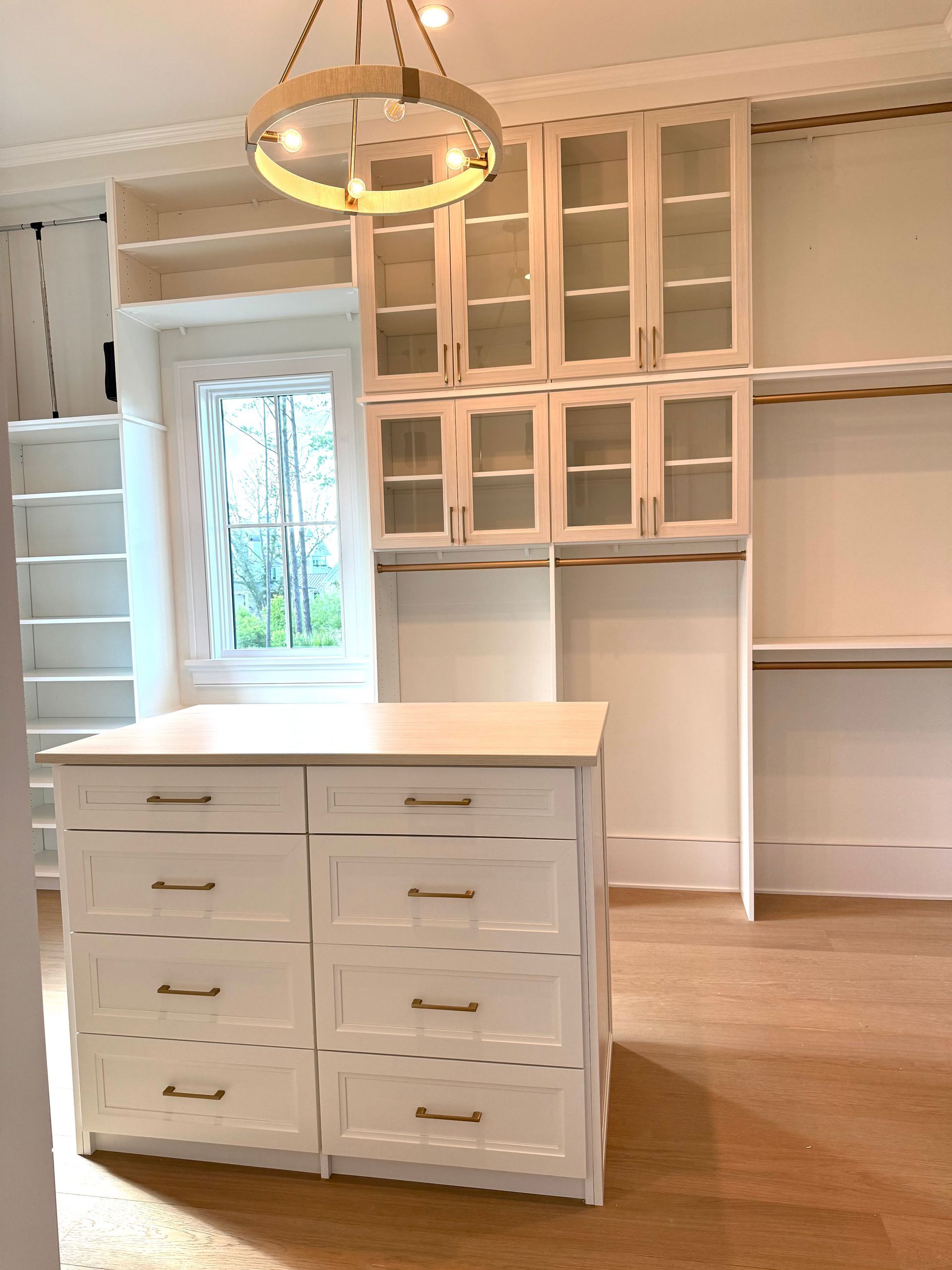 White walk-in closet with island dresser, glass-front cabinets, and a window.