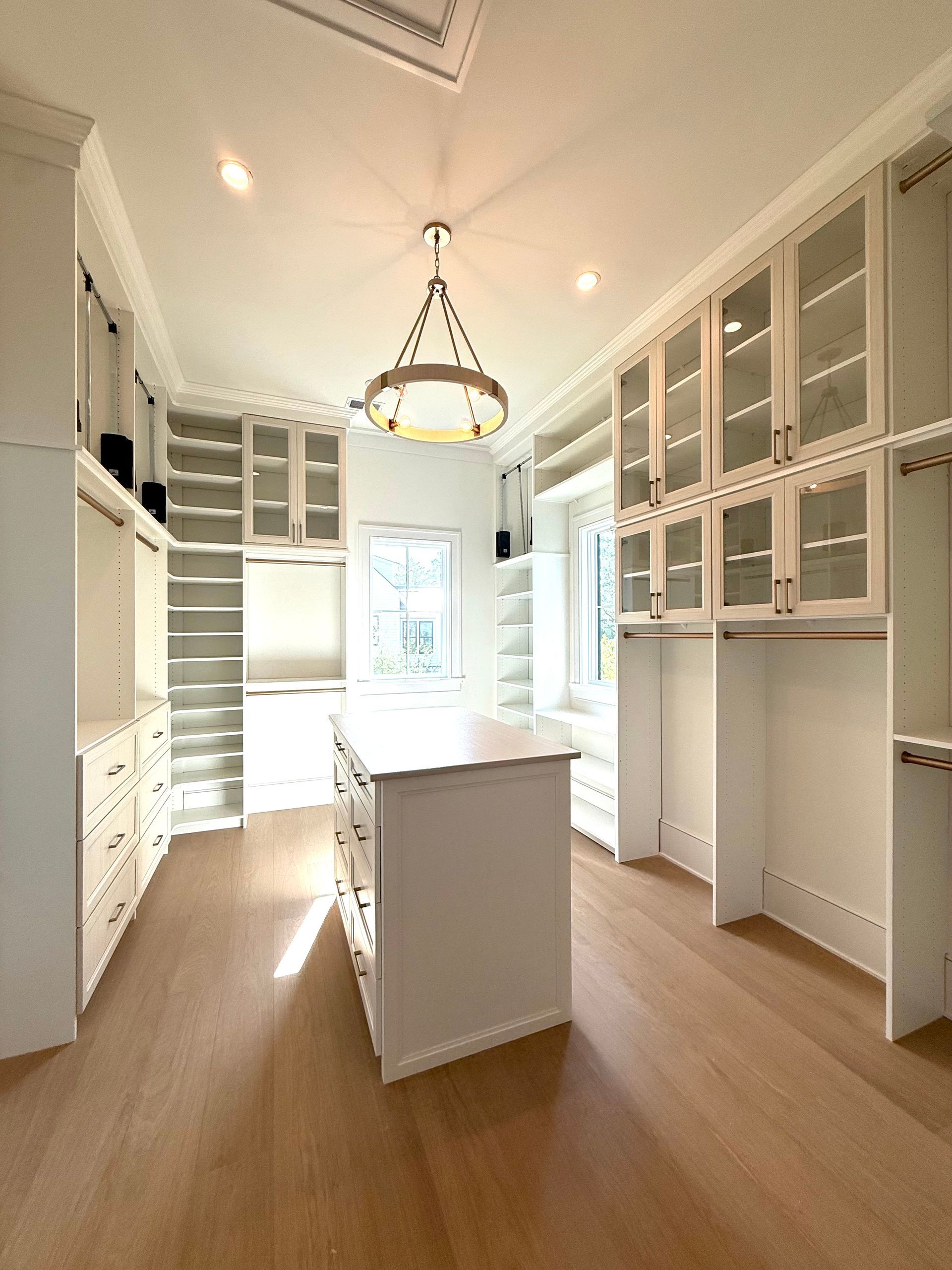 Spacious, white walk-in closet with island, glass-door cabinets, and light wood flooring.