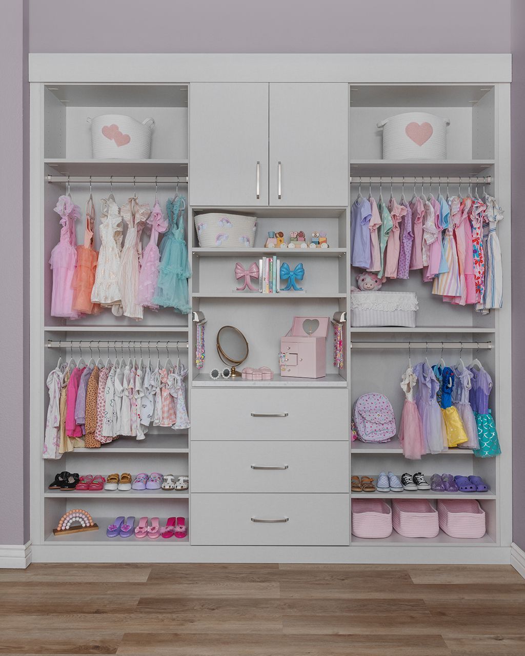 White built-in closet with pastel children’s clothes, shelves, drawers, and pink storage bins on a wood floor.