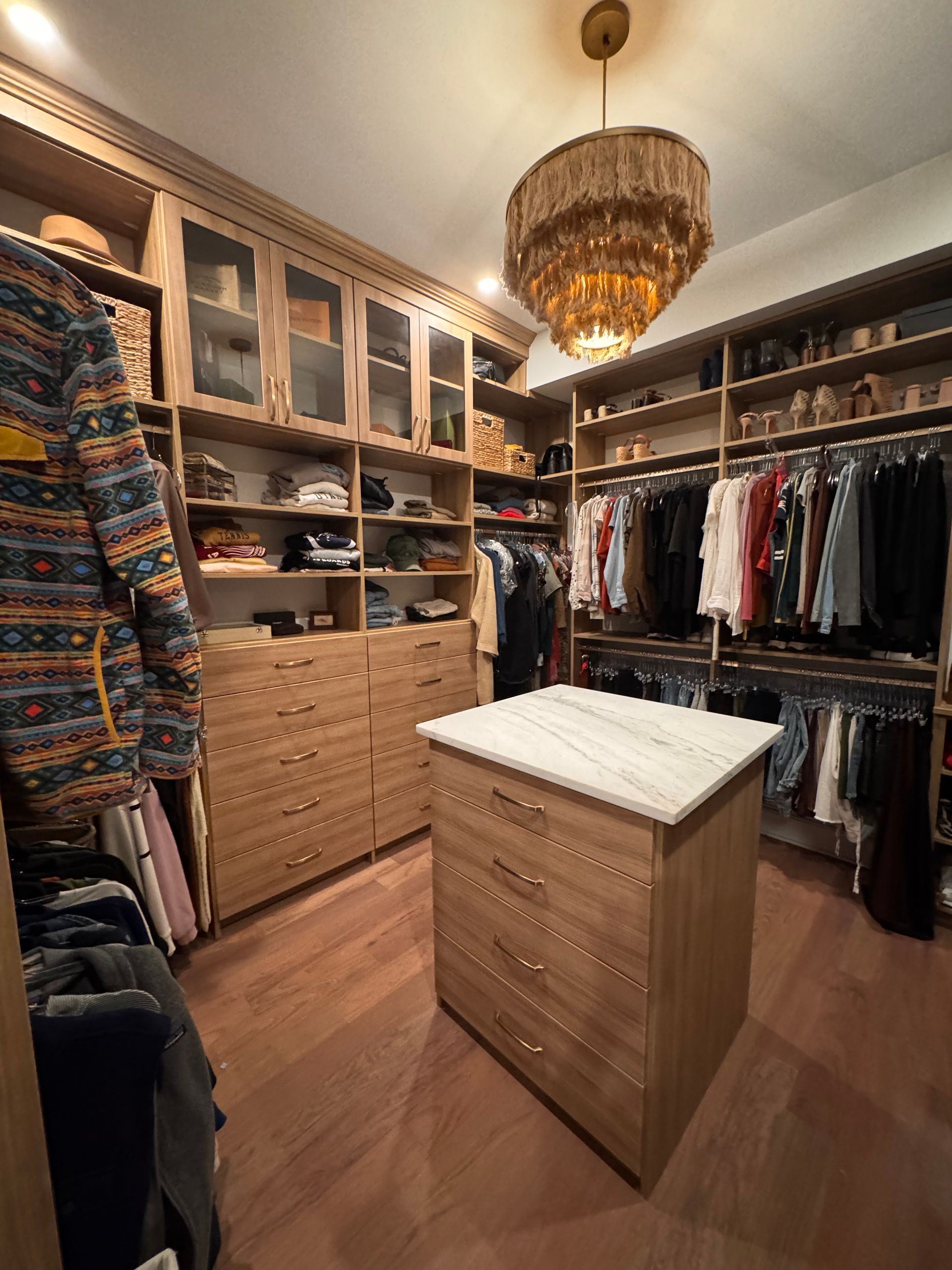 Walk-in closet with light wood shelving, drawers, and an island with a marble top. Clothes hang on racks.