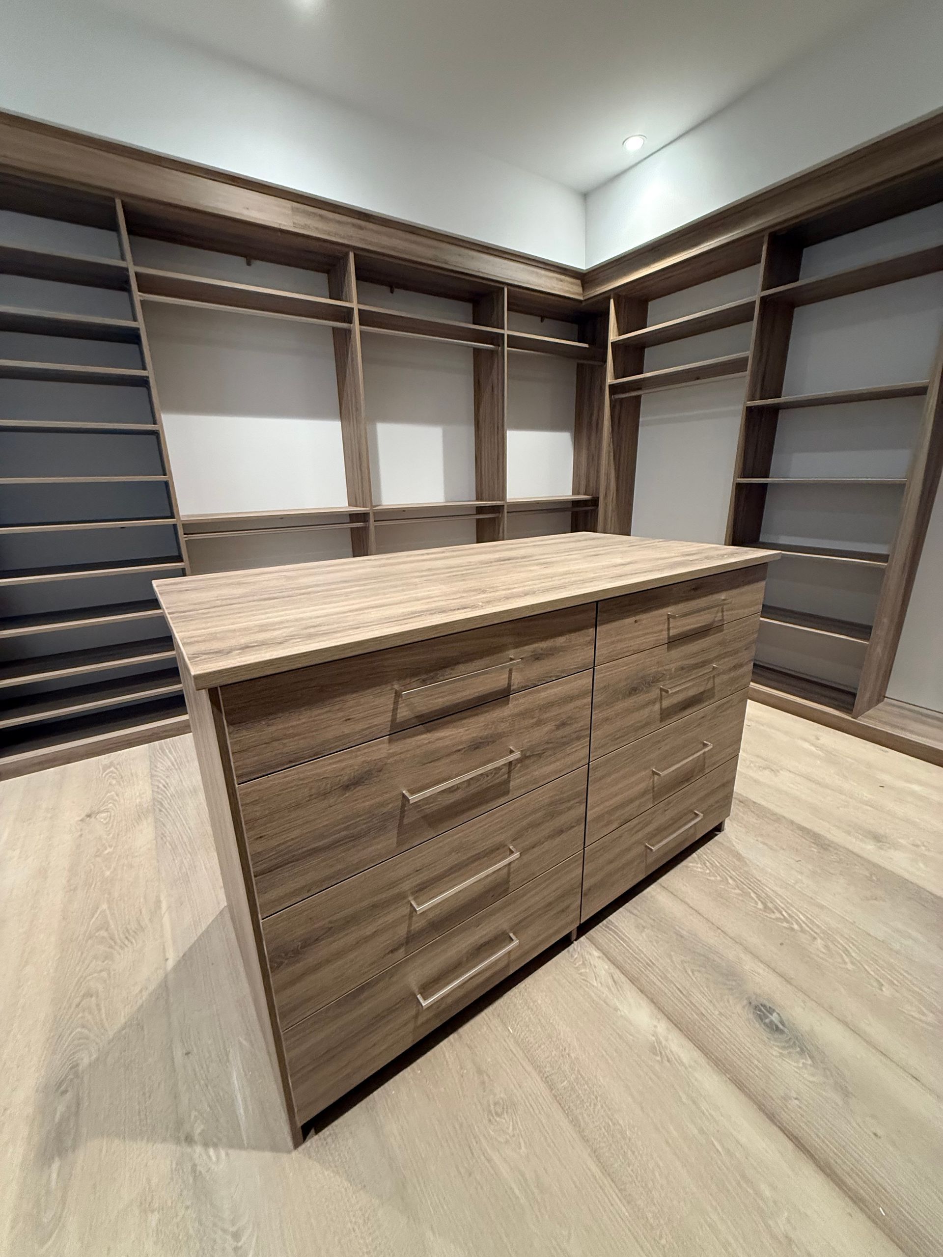 Walk-in closet with built-in shelves and a central dresser. Light wood and neutral colors create a minimalist space.