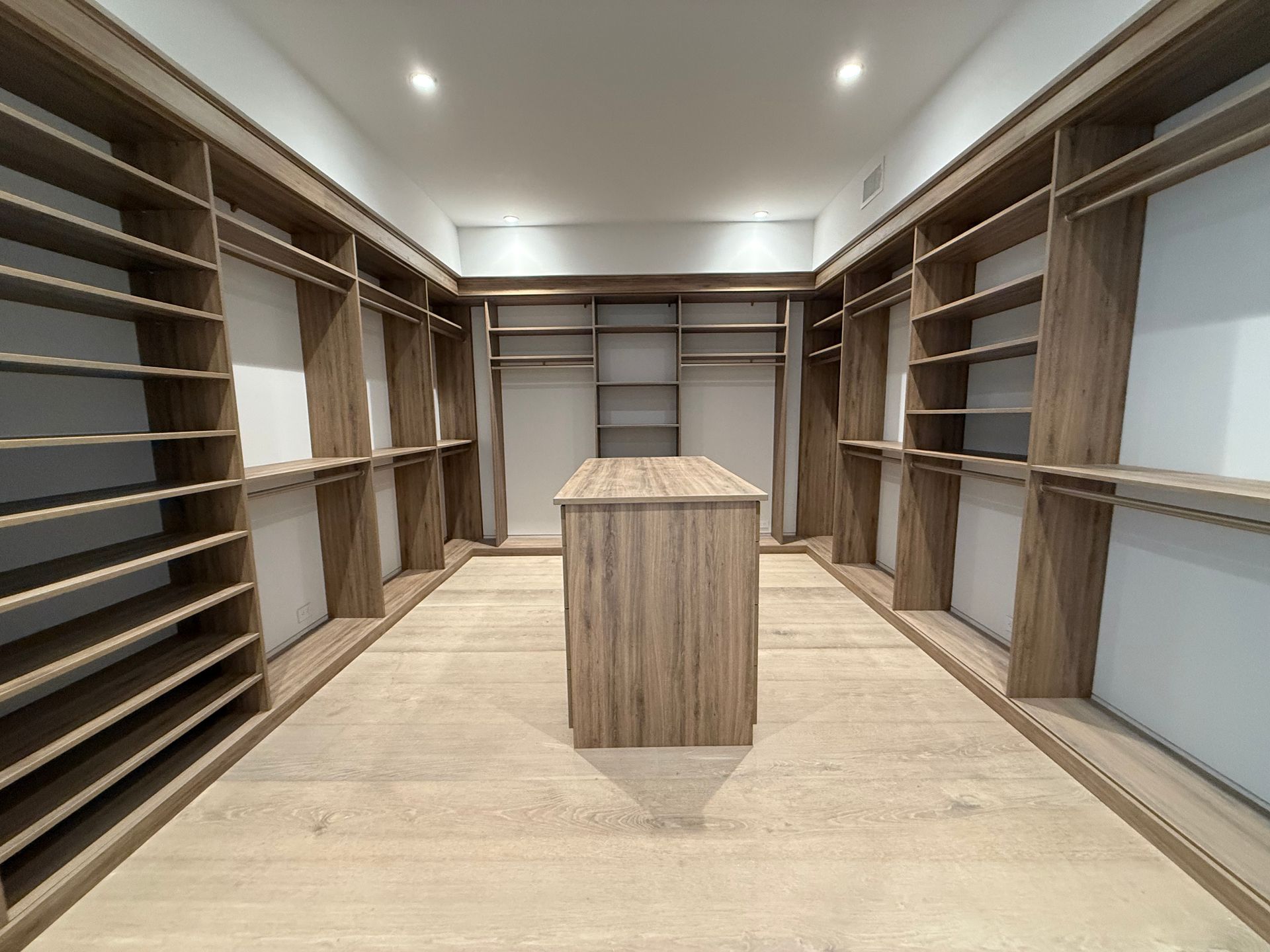 Empty walk-in closet with built-in shelves and a central island. Light wood, neutral tones, and recessed lighting.