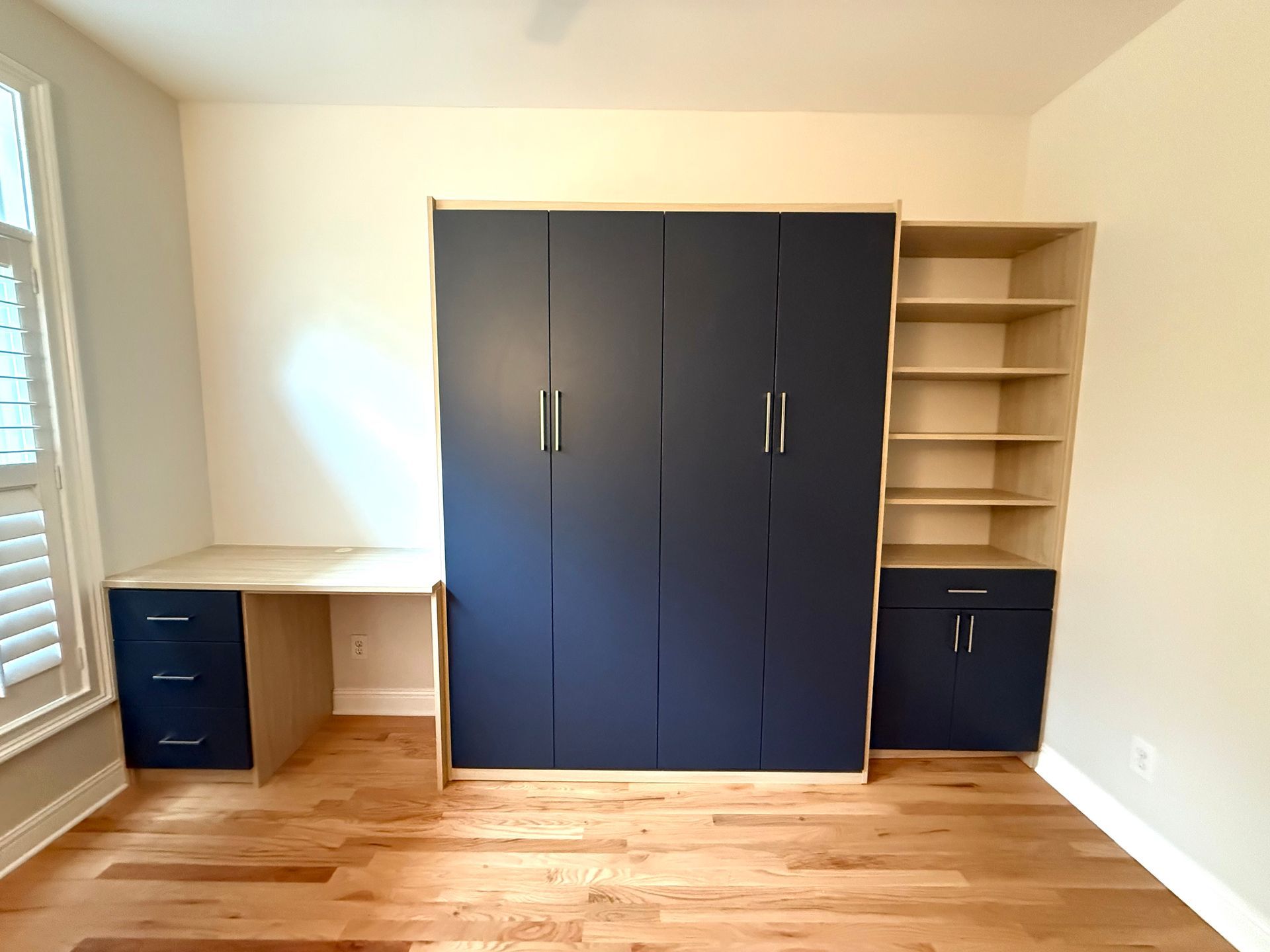 Built-in navy blue wall bed, desk, and bookshelf unit in a room with wood floors and a window.