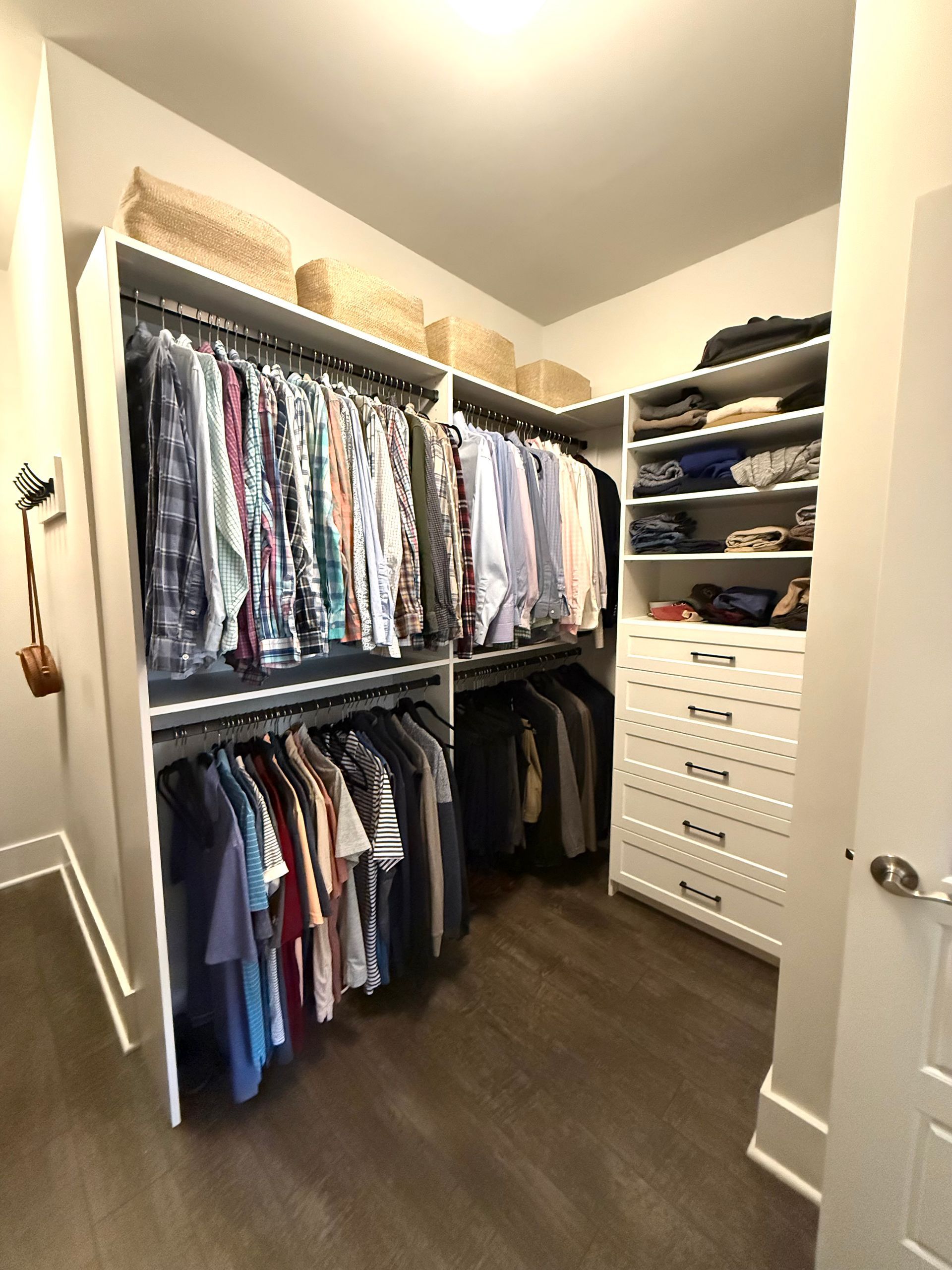 A walk-in closet with hanging clothes, shelves, and drawers. White shelving against brown floors and a beige wall.