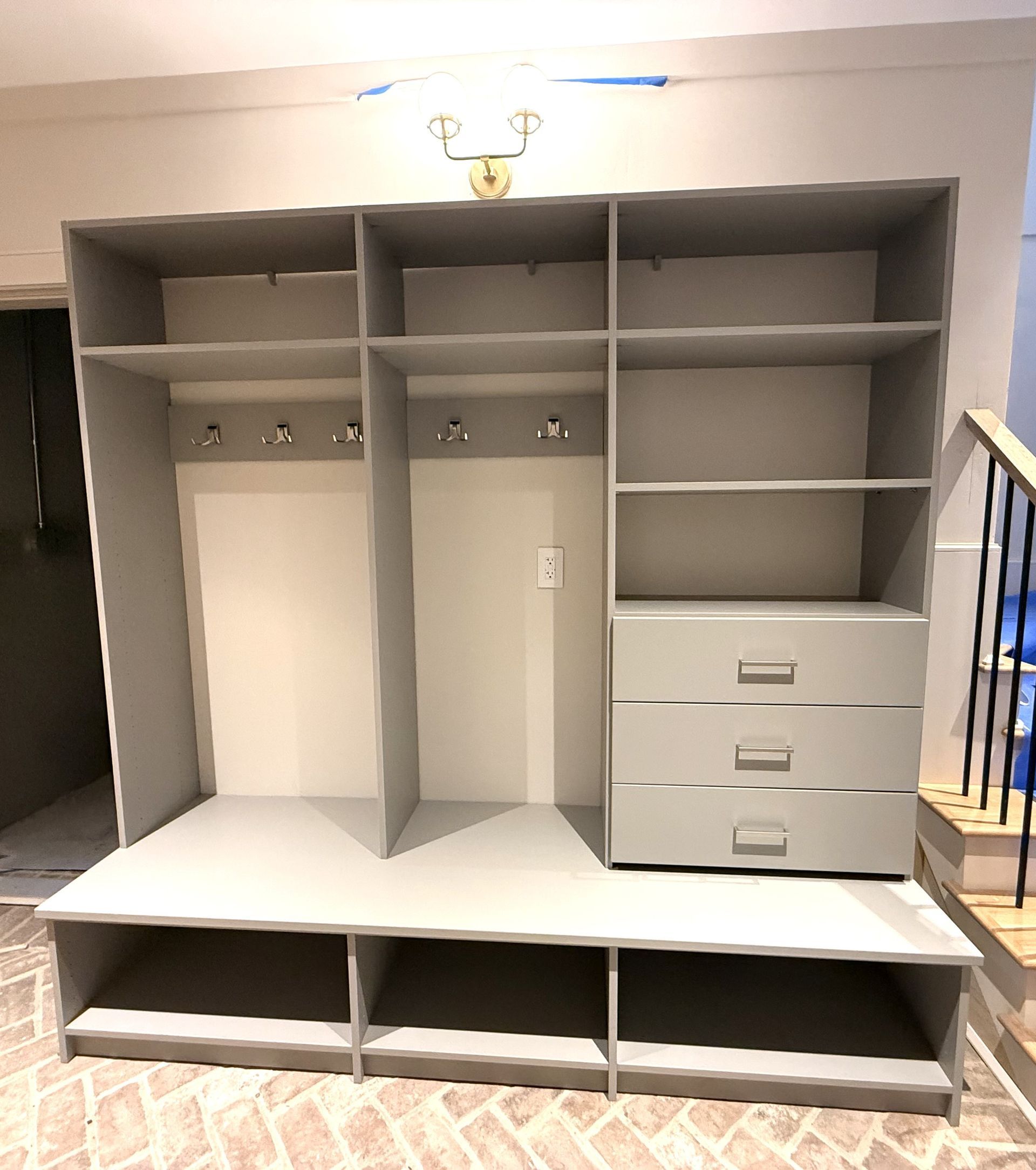 Grey built-in storage unit with shelves, hooks, drawers, and lower cubbies, on a brick floor.
