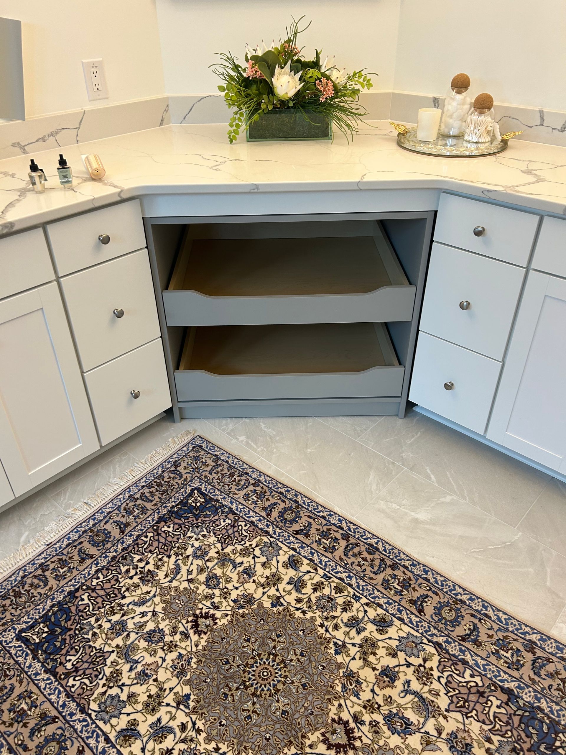 Kitchen with white cabinets, marble countertop, and open drawers. A floral arrangement and rug are in view.