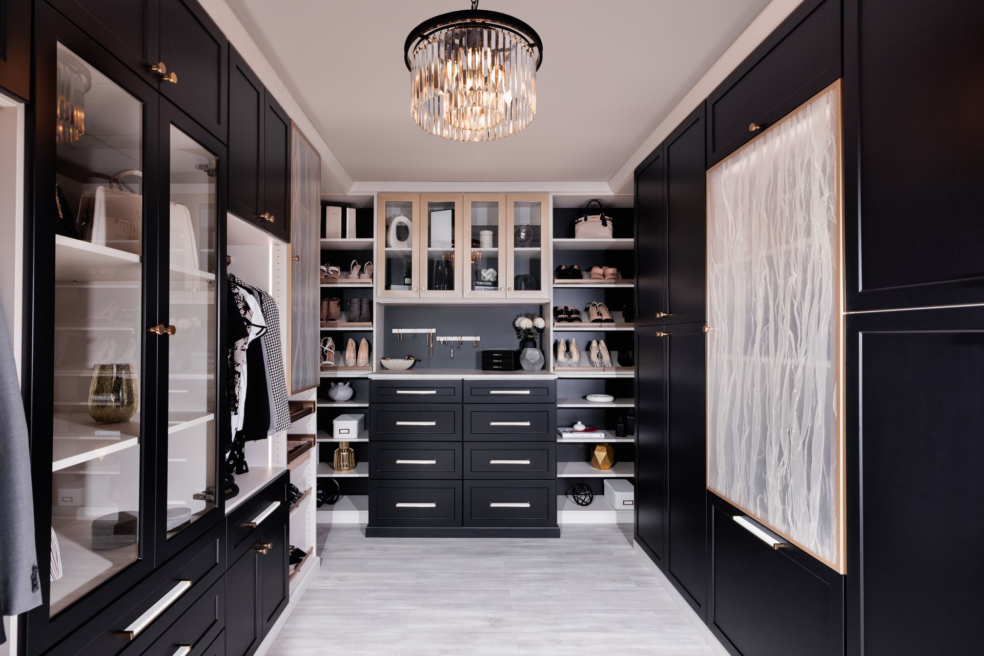 Elegant black walk-in closet with glass cabinets, drawers, and a crystal chandelier