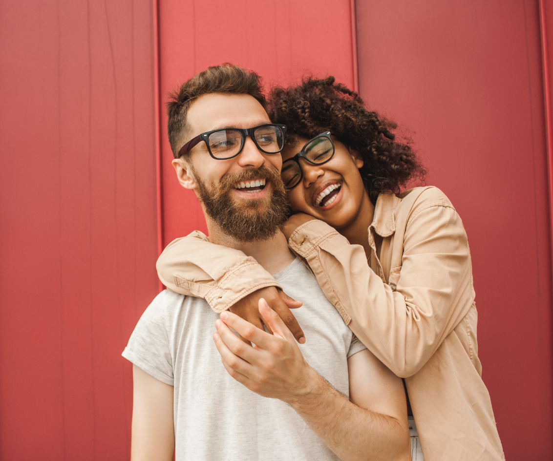 A man and a woman are hugging each other in front of a red wall.