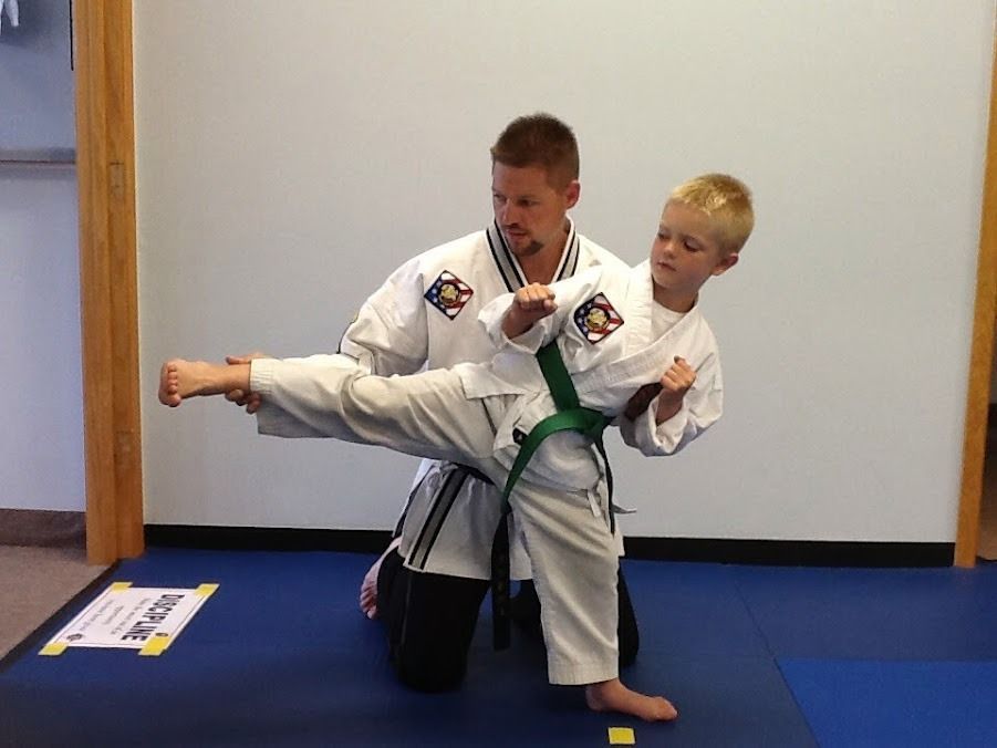 An instructor in a martial arts uniform kneels and supports a student in a side kick position on a blue gym mat.