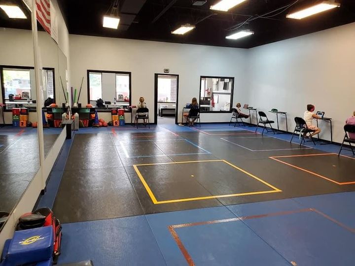 A spacious gym with mats featuring colorful square tape outlines, occupied by several people seated at individual desks.