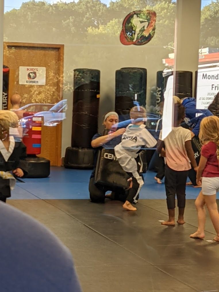 A martial arts instructor holds a target pad for a student in a white uniform while other children wait in a gym.