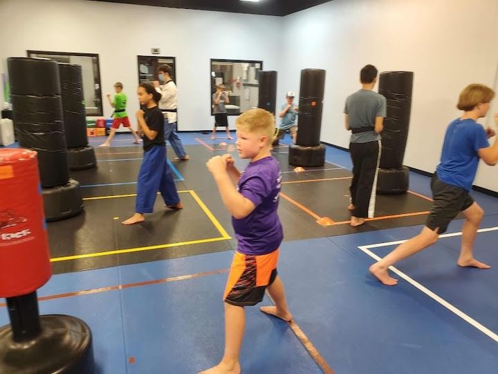 Students in a martial arts class practice fighting stances in front of standing punching bags on a gym mat.