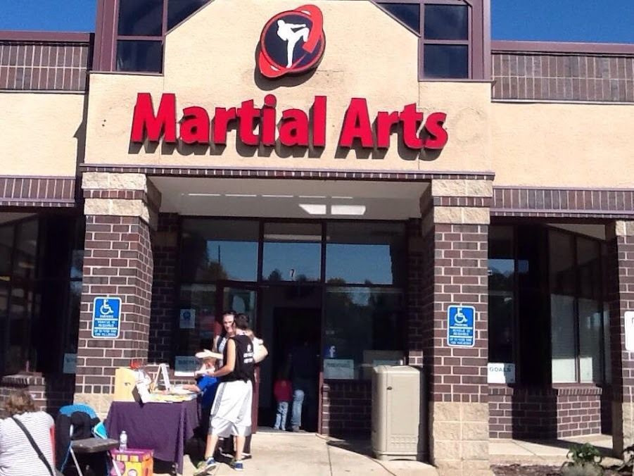 Exterior of a martial arts studio featuring a red sign, brick pillars, and people standing by an outdoor table.