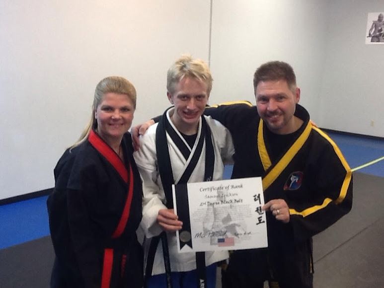 Three people in martial arts uniforms stand together, with the center person holding a black belt rank certificate.