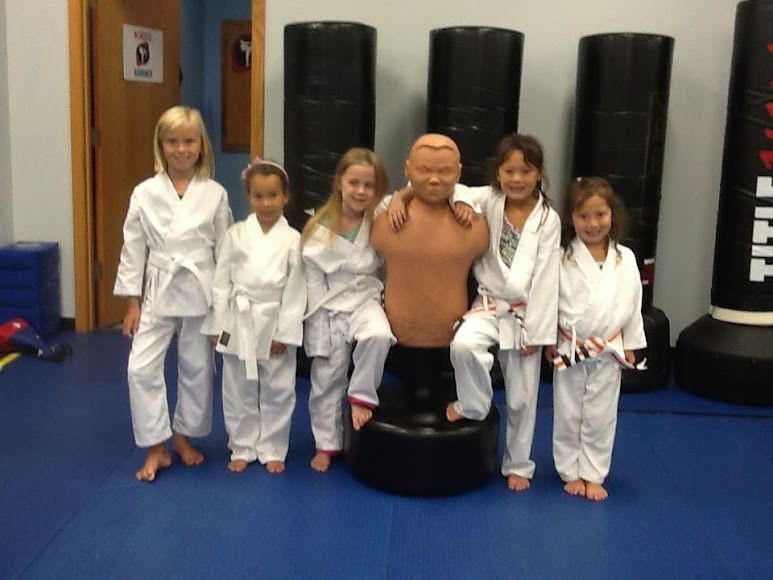 Six students in white martial arts uniforms pose around a training dummy in a gym with blue flooring and tall punching bags.