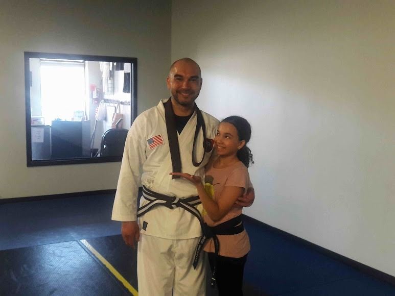 A person in a white martial arts uniform with a black belt poses with a person wearing a black belt over a pink shirt.