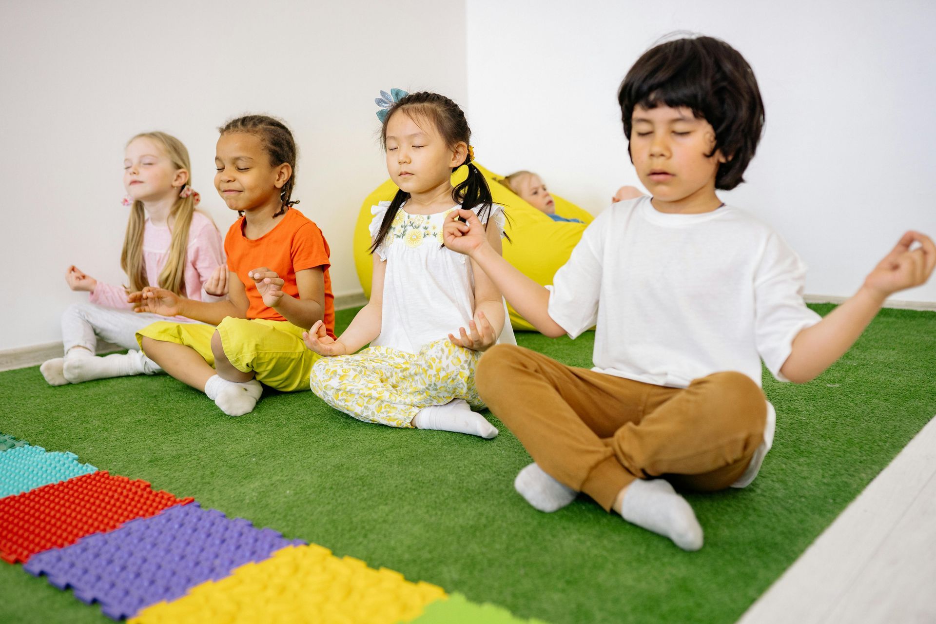 Children sitting cross-legged on green turf, meditating with their eyes closed in a brightly lit room.