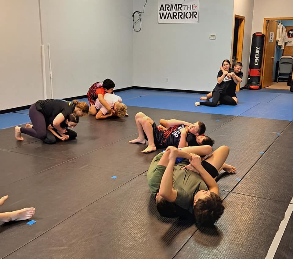 Students practice Brazilian Jiu-Jitsu grappling techniques on mats in a training studio.