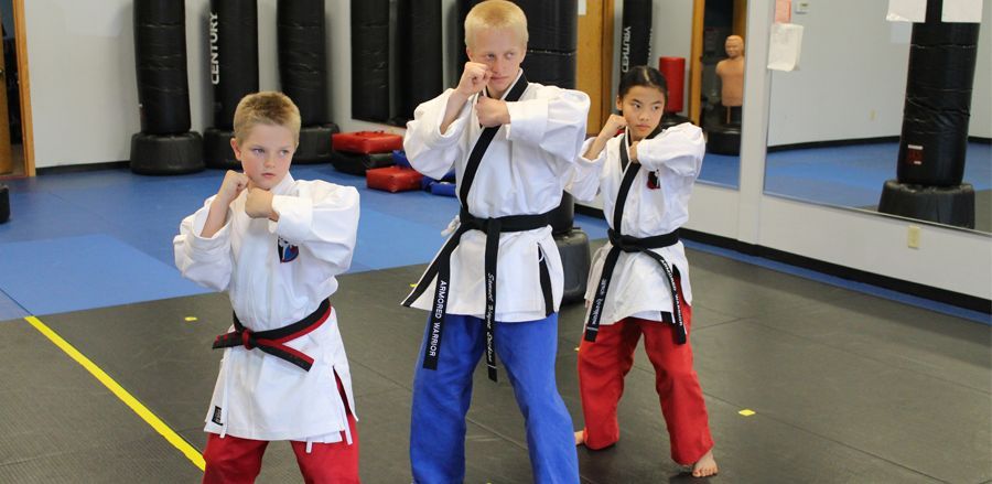 Three students in white martial arts uniforms and black belts practice defensive stances in a training studio.