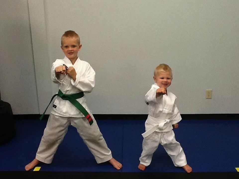 Two children in white karate uniforms perform a punch pose on a blue mat against a plain wall.