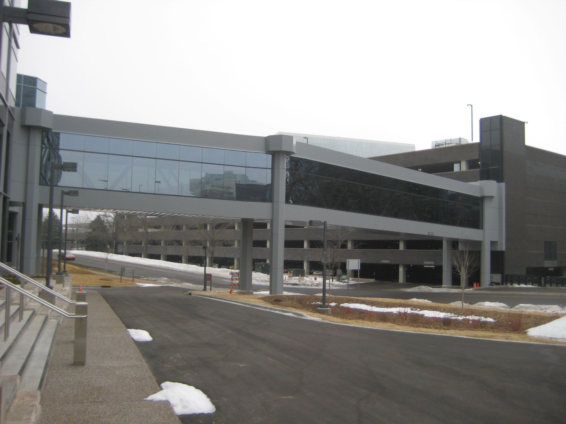 Skywalk connecting two modern buildings, over a parking garage. Gray and glass construction; snowy ground.