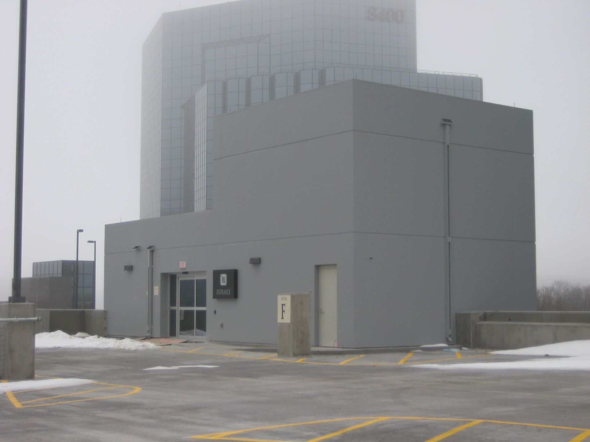 Gray building entrance on a parking deck, with a skyscraper in the background on a foggy day.