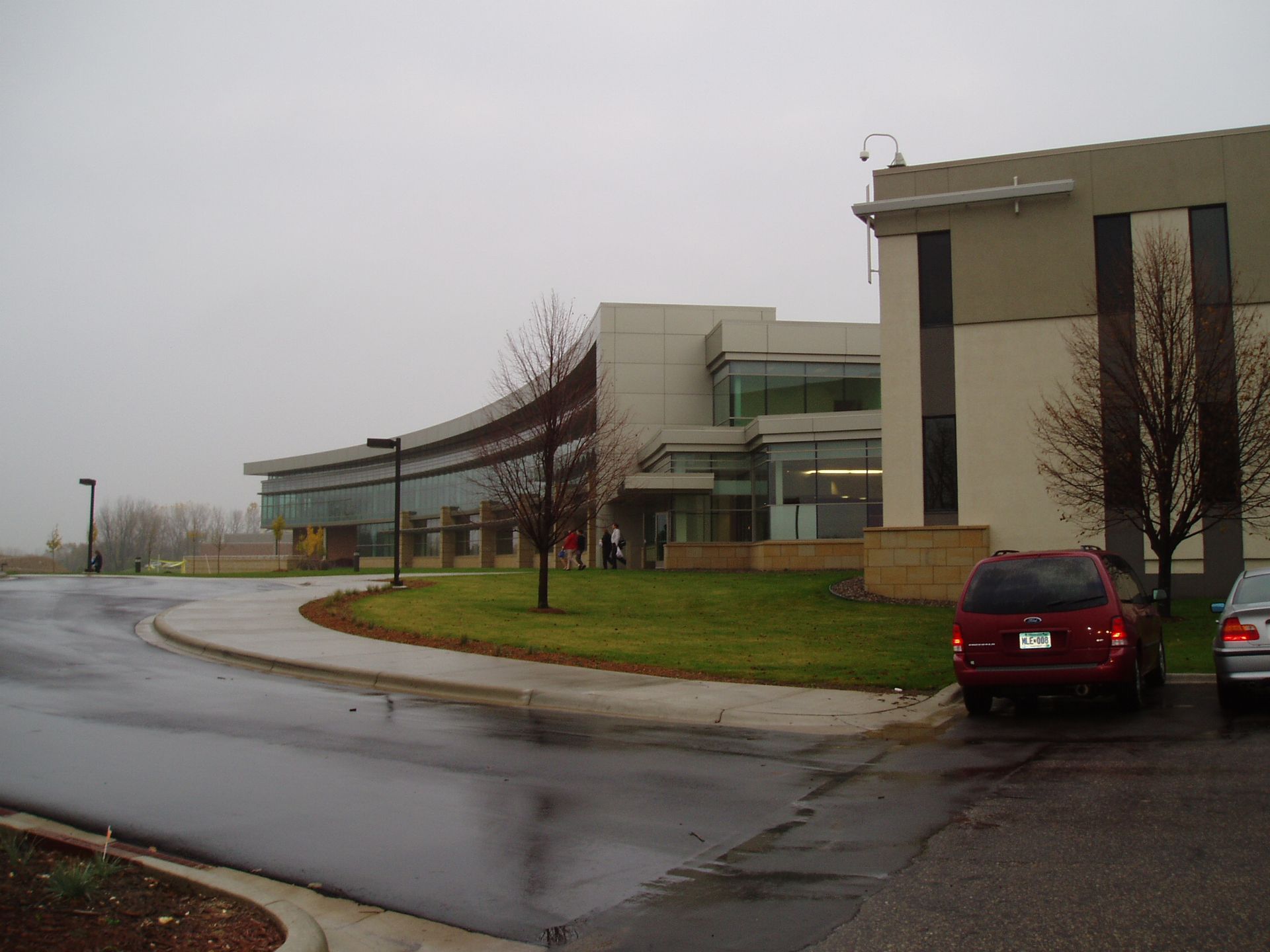 Modern building with curved glass facade, driveway, parked cars, and overcast sky.