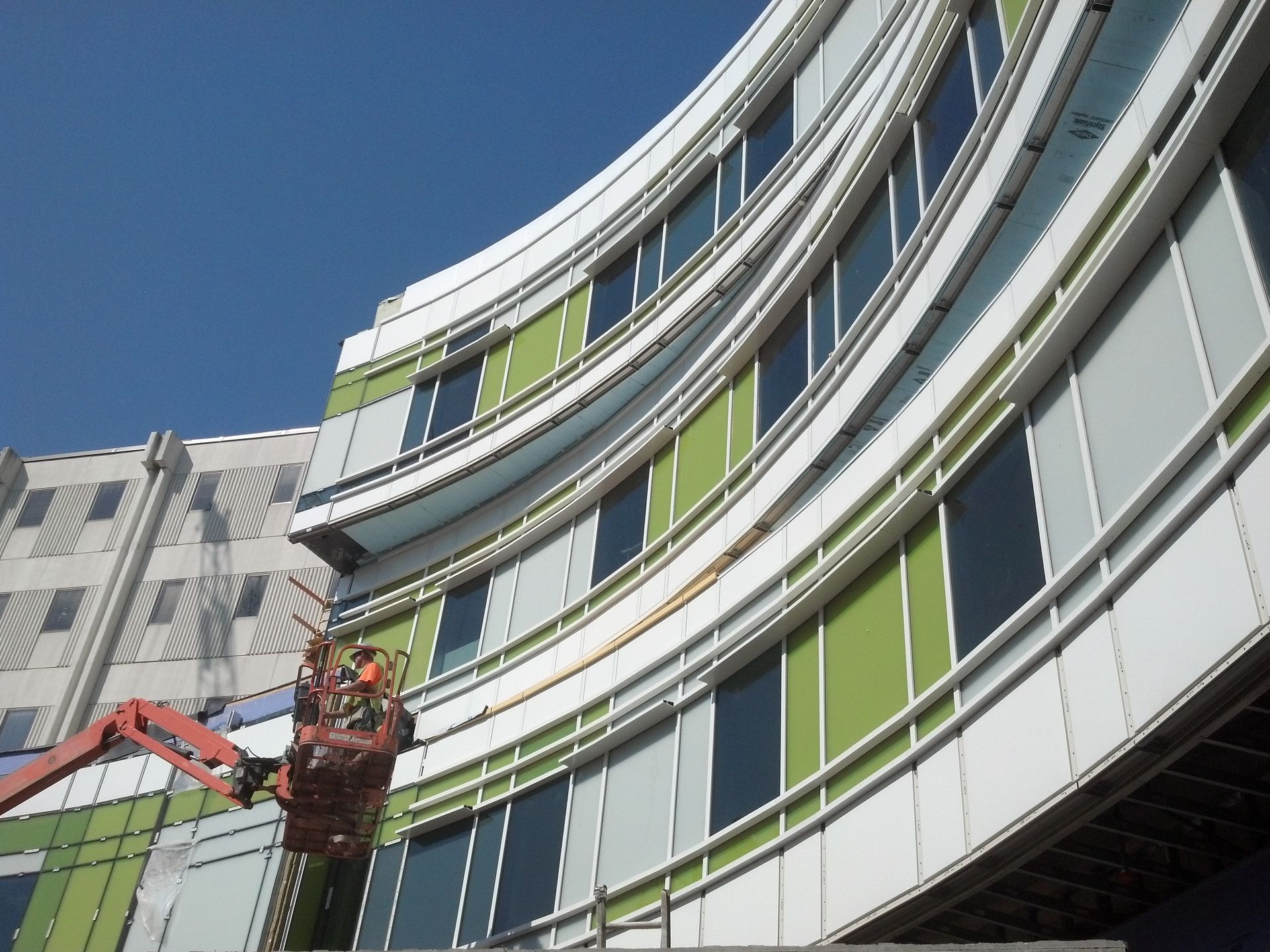 Curved building with green and white panels, construction worker in lift repairing exterior under a blue sky.
