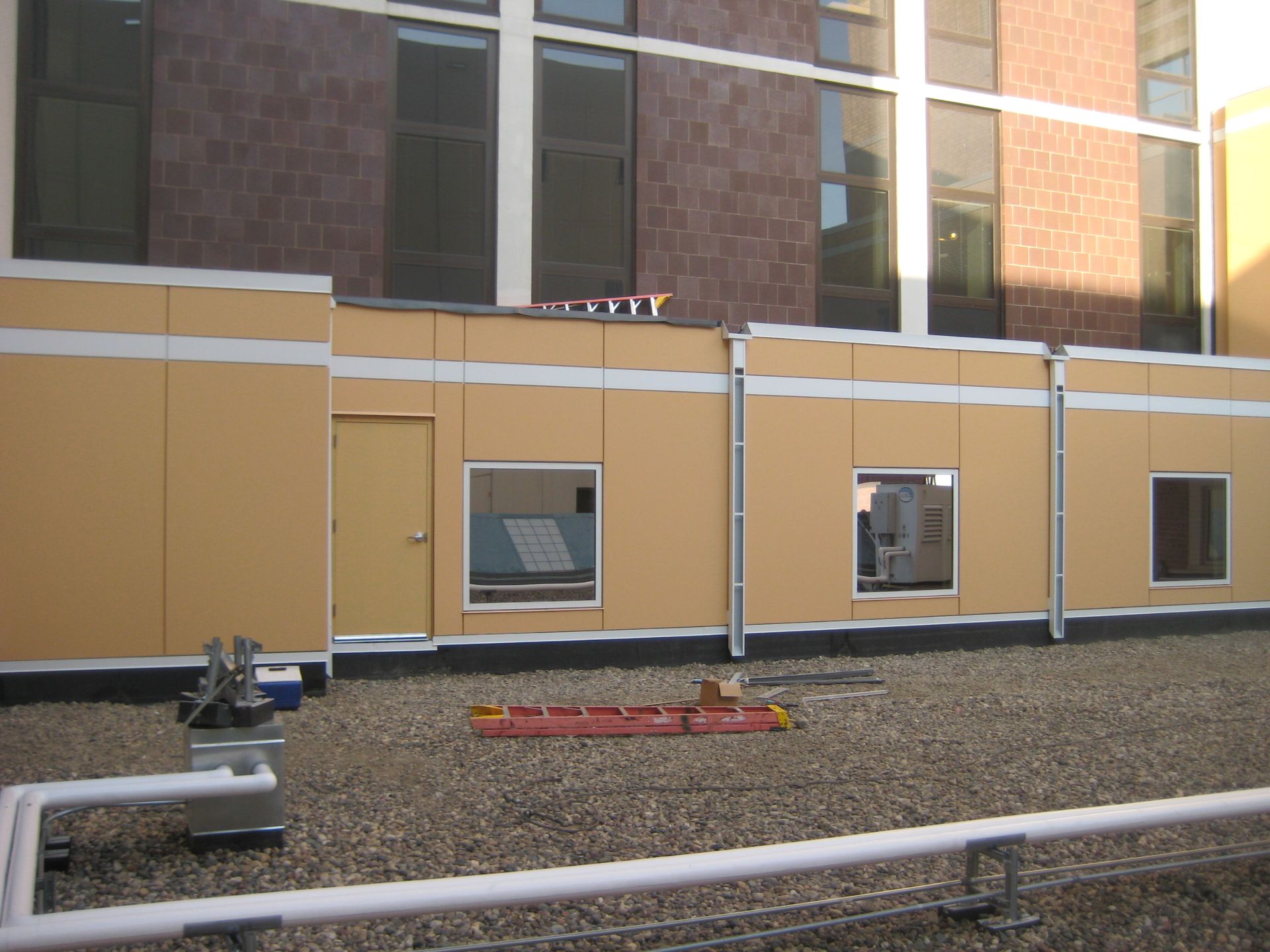 Yellow rectangular structure with windows and a door on a gravel rooftop against a building facade.