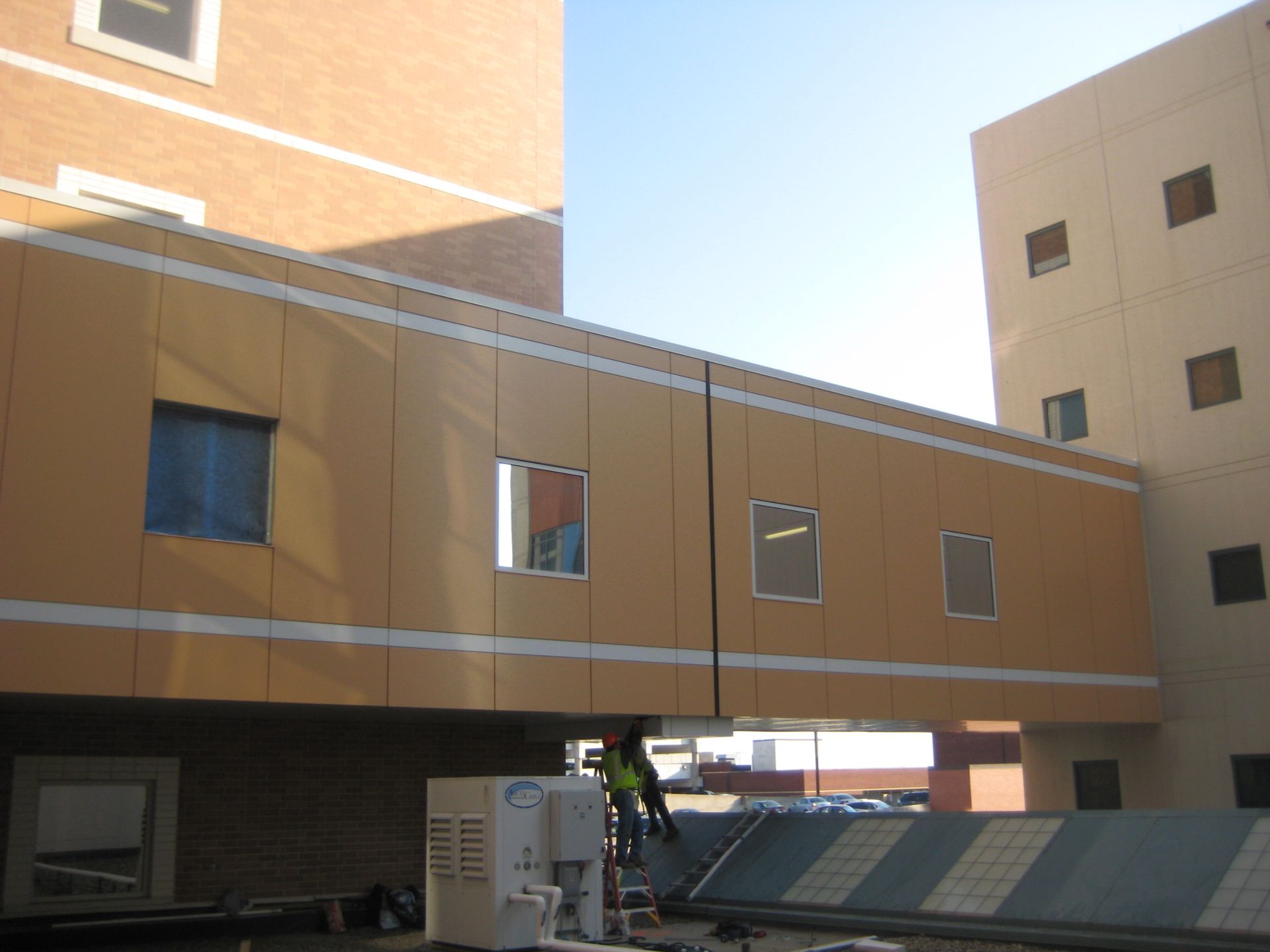 Exterior view of a beige and brown bridge connecting two buildings. A person is visible below, near the roof.