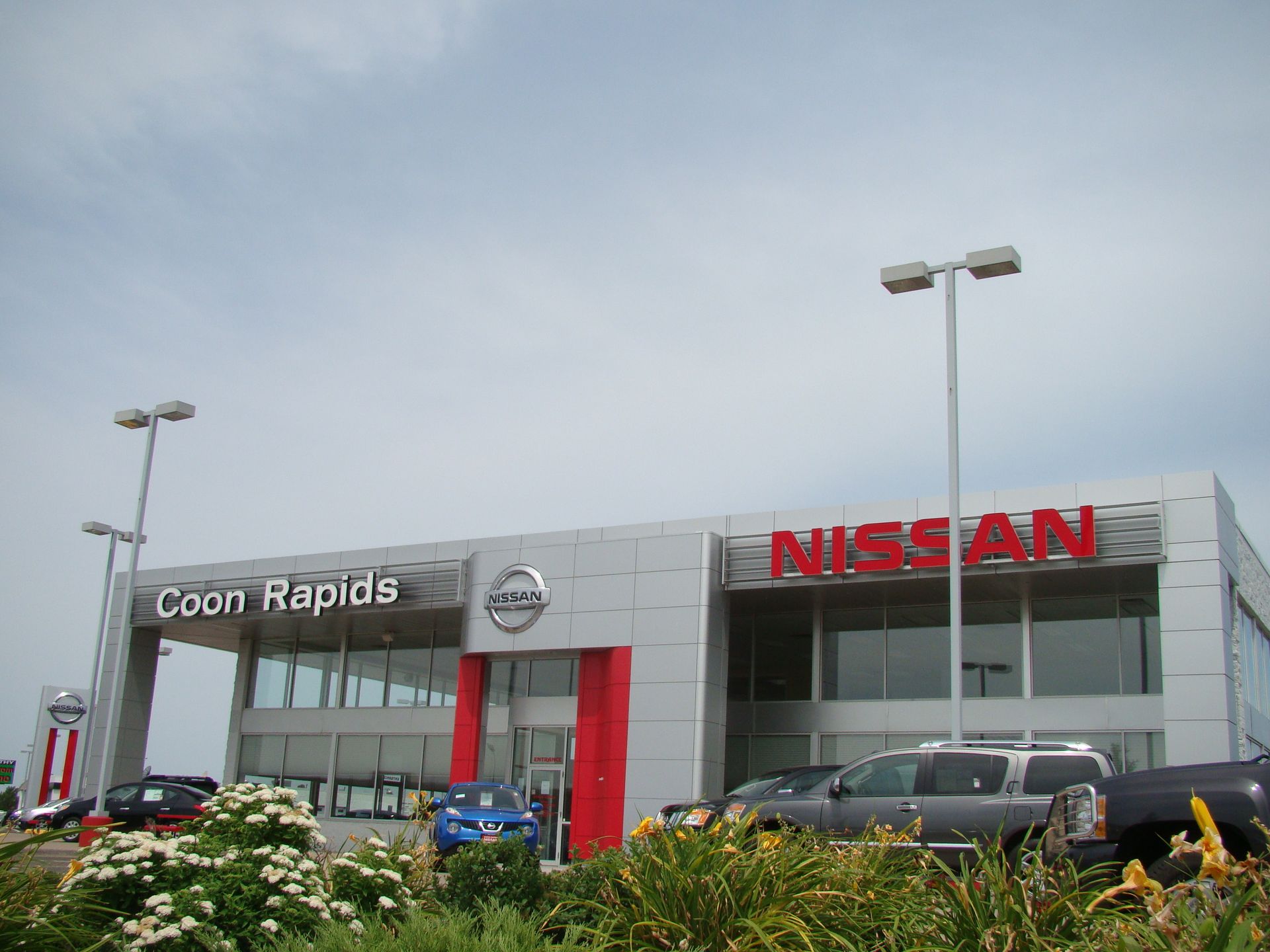 Coon Rapids Nissan dealership with cars in front. Gray building with red accents, 