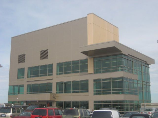 Tan, three-story building with large windows and a flat roof, over a parking lot with cars.